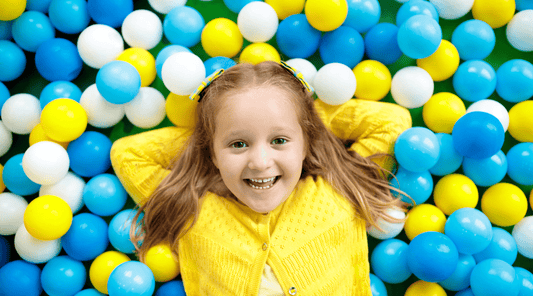 A young child smiling and lying down on a floor filled with colorful balls, at a play area.